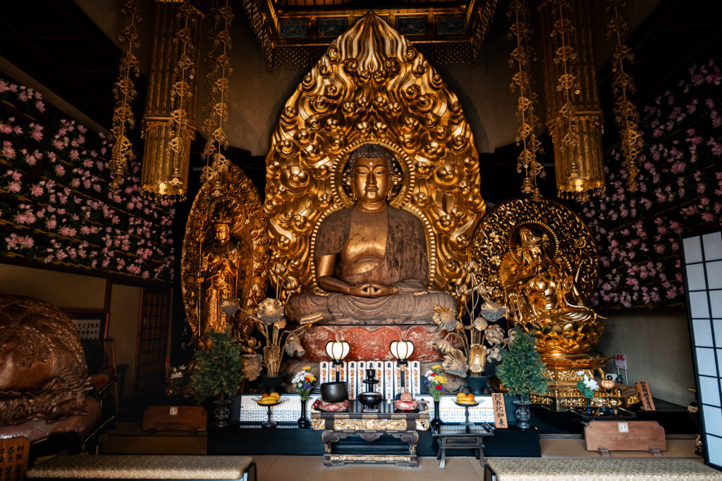 Golden Amida Buddha statue inside Hasedera Temple hall in Kamakura, surrounded by ornate gilded decorations and floral offerings.