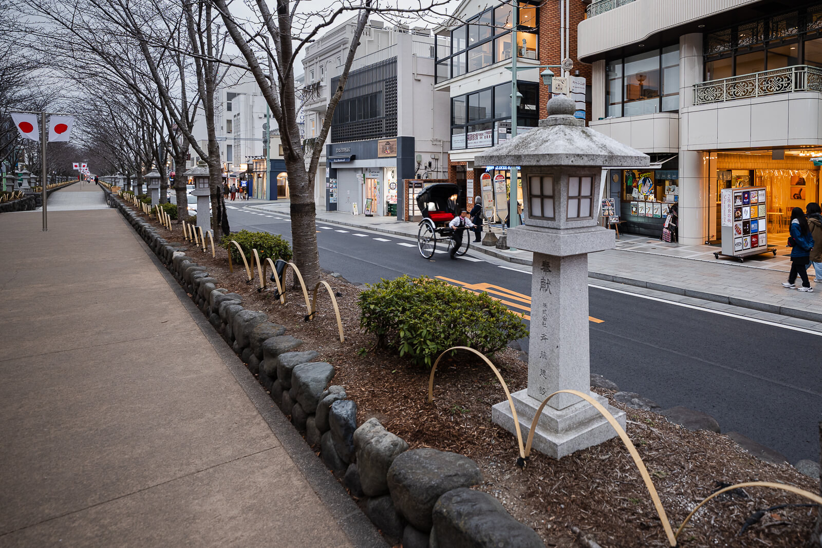 Stone lantern beside the raised Dankazura promenade with shops and bare cherry trees in central Kamakura