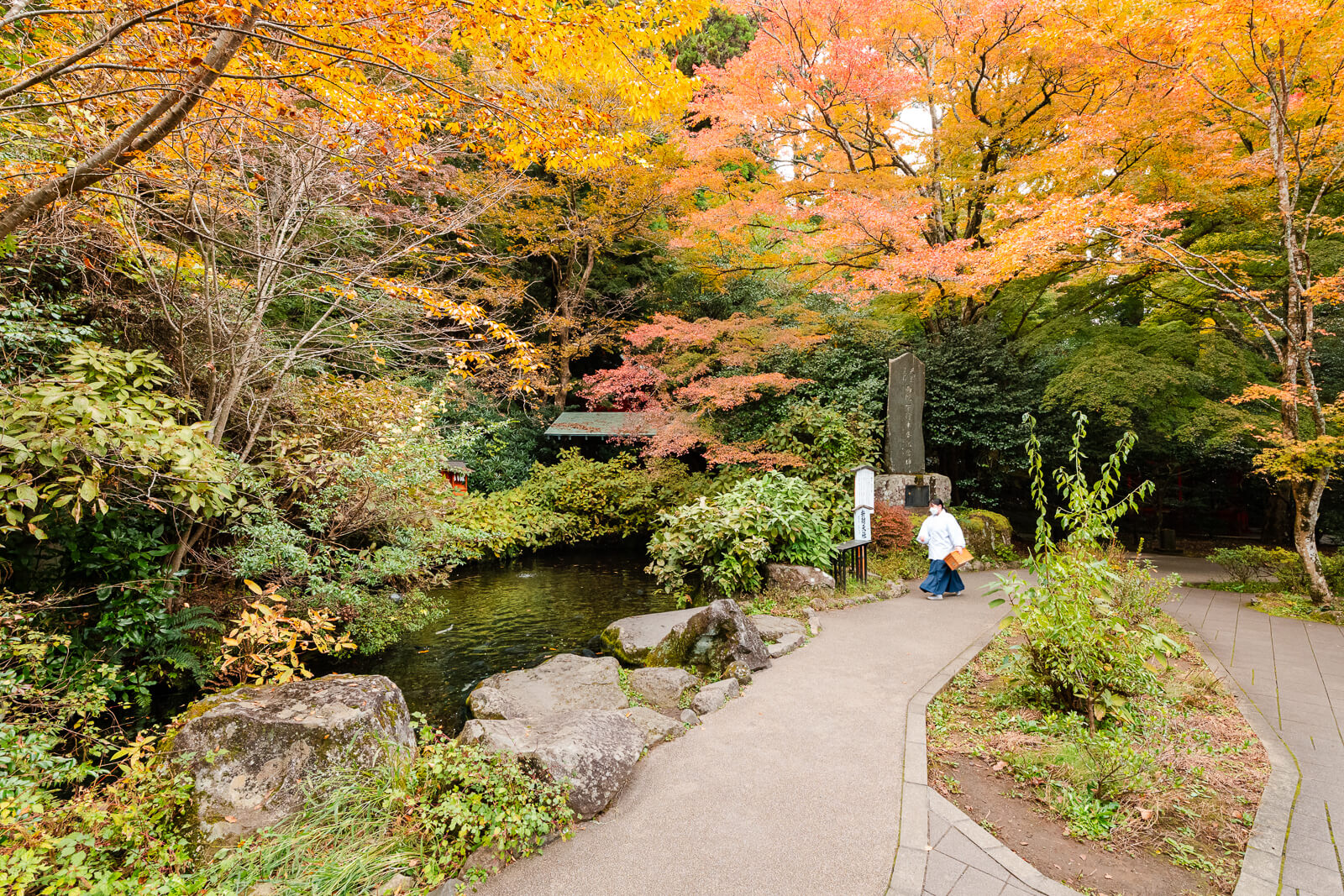 Visitors on a curving pond path beneath orange and yellow autumn foliage at Hakone Shrine in Motohakone