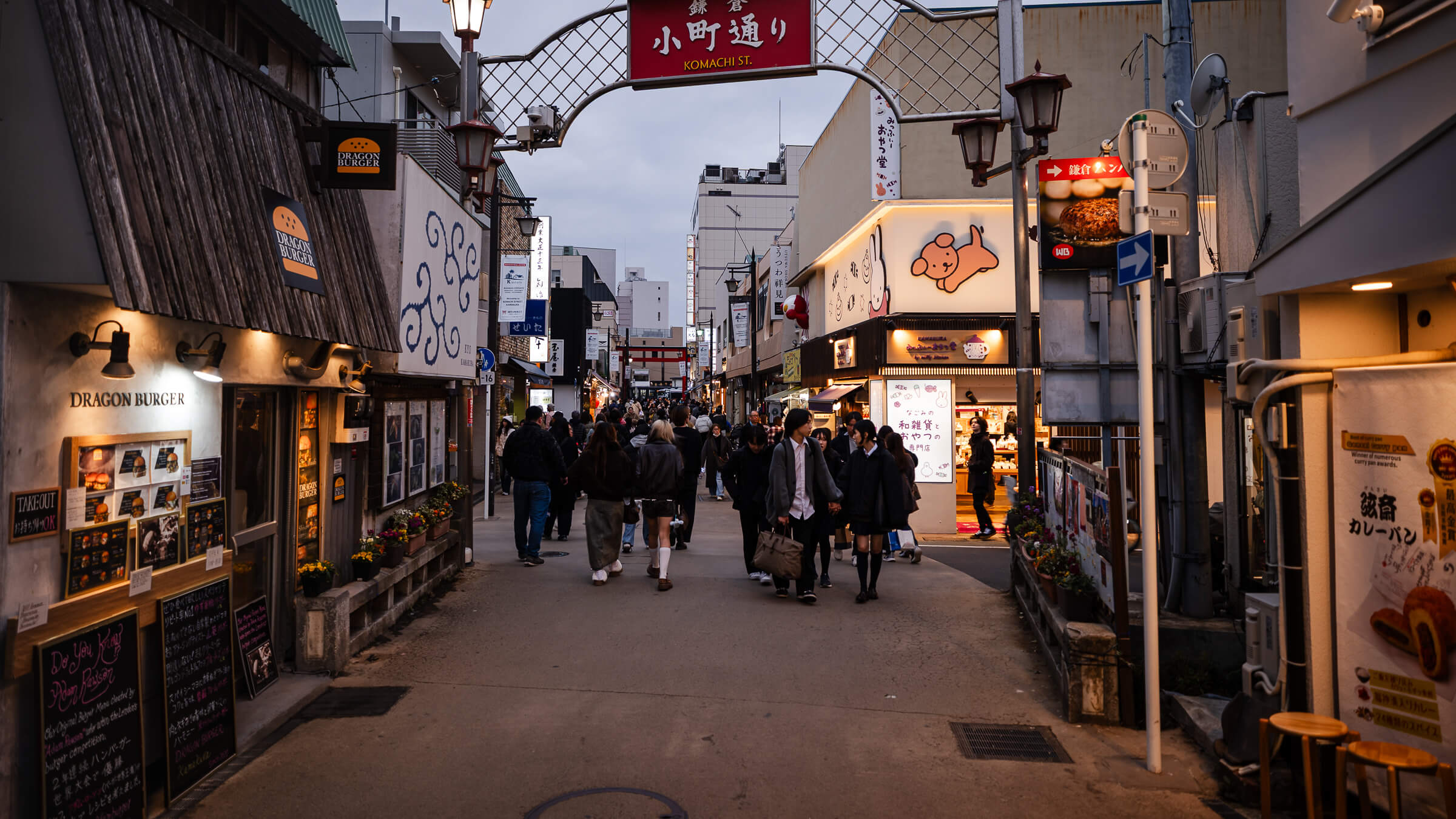 Komachi-dōri entrance in Kamakura at dusk with lanterns, shops, and evening crowds walking toward Tsurugaoka Hachimangū