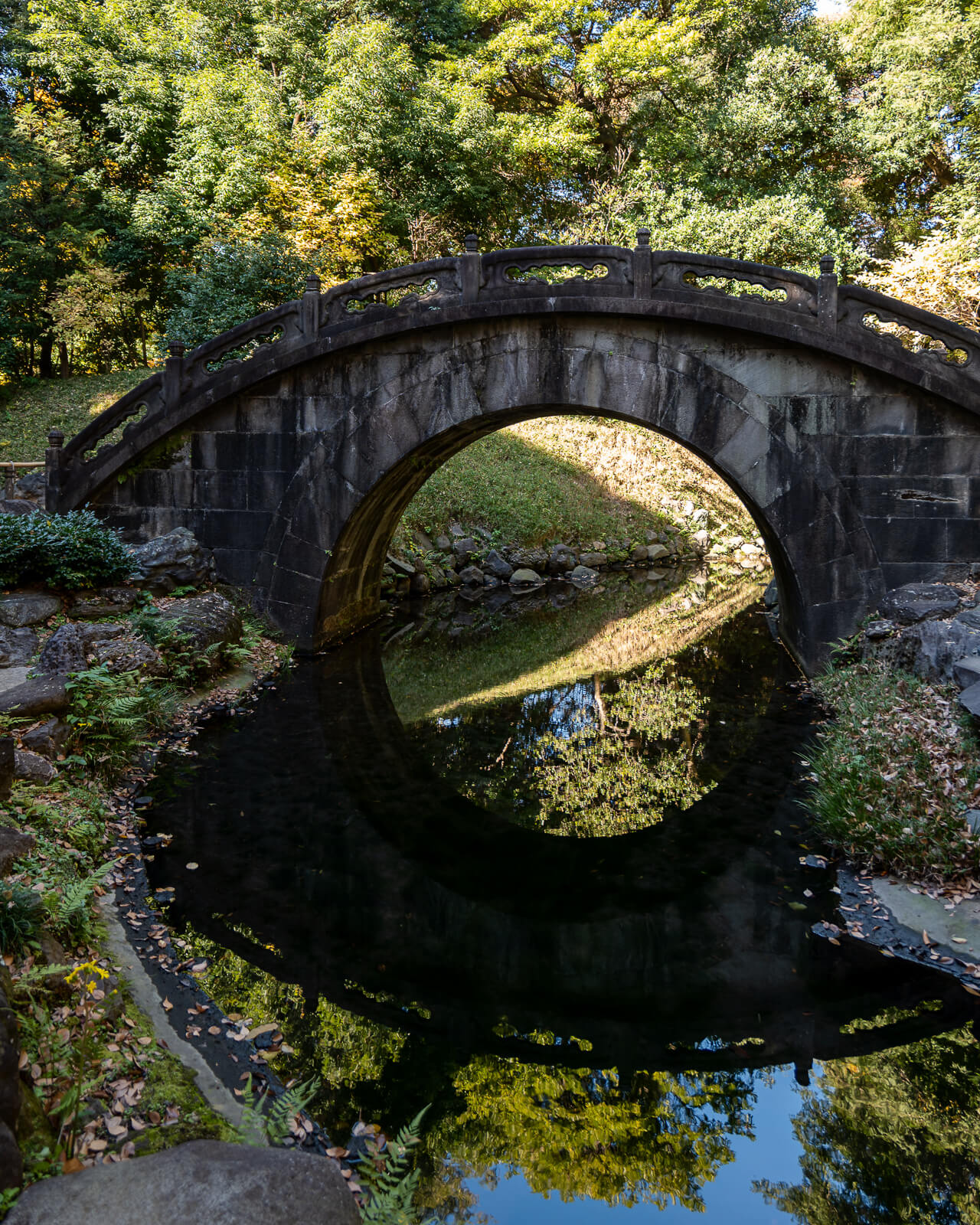 A stone arch bridge crosses a narrow waterway at Koishikawa Korakuen, with its curved shape reflected beneath autumn trees.